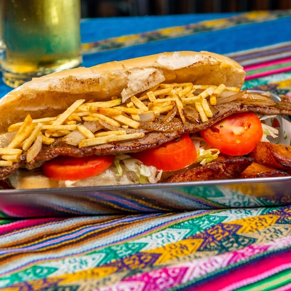 A Latin-American steak sandwich with fries, tomatoes, and lettuce on a colorful patterned tablecloth.