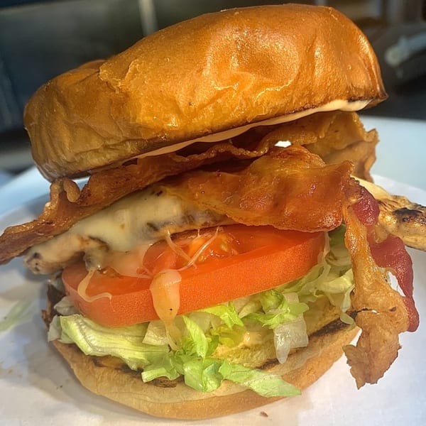 A close-up of a chicken sandwich with melted cheese, bacon, tomato, and lettuce on a bun.