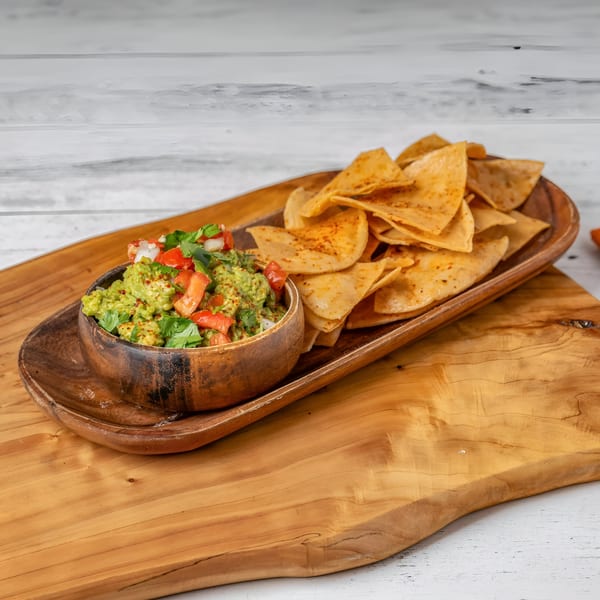 Guacamole with tomatoes, onions, and cilantro, served with tortilla chips on a wooden platter.